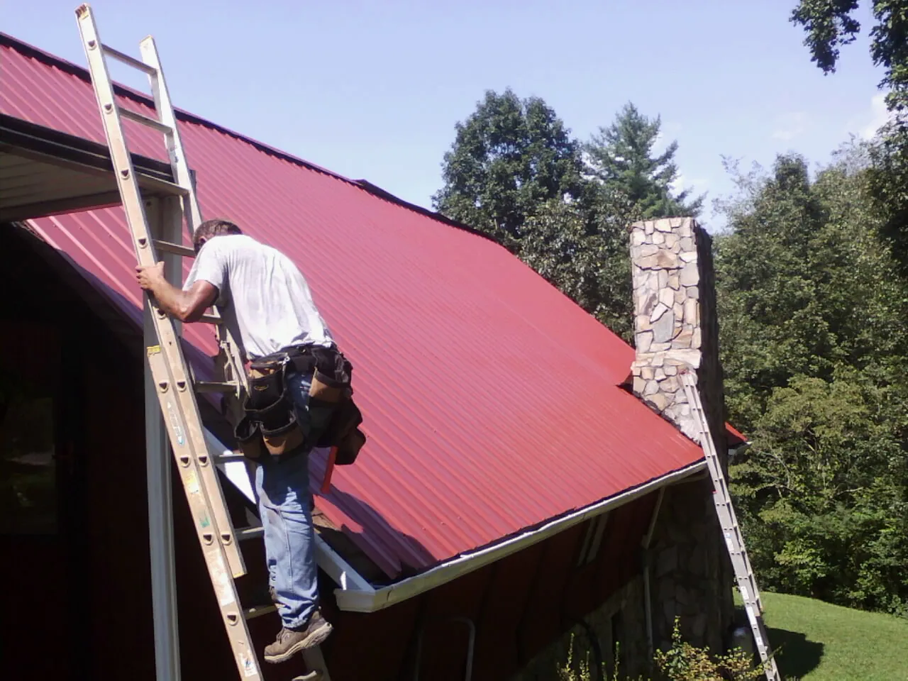 Roofer on a ladder beside a red metal roof and stone chimney — Soffit & Fascia Repair by Roofing Brothers in West Yarmouth