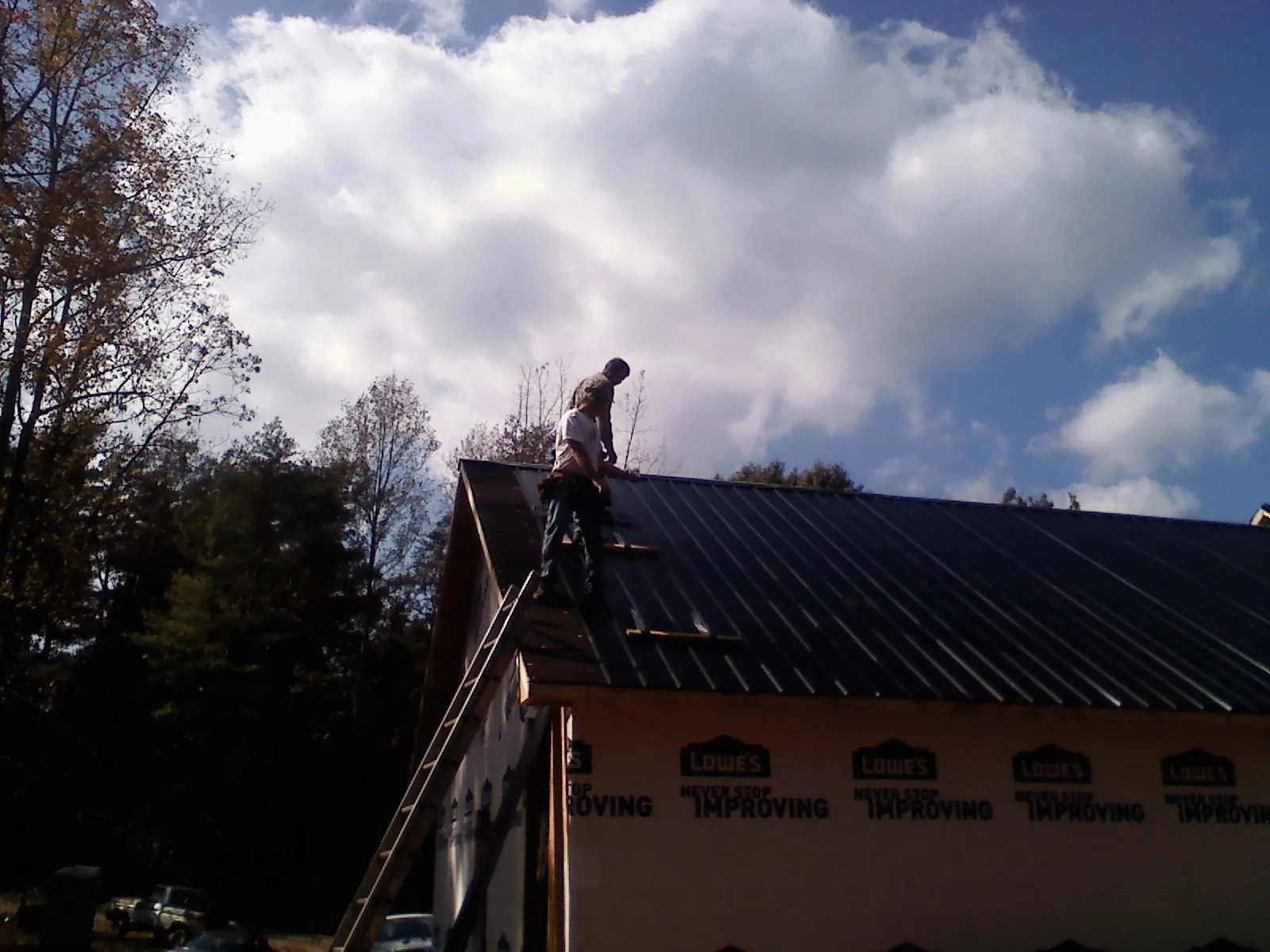 Roofer fastening dark metal panels high against a cloudy sky, from a Roof Inspection job in West Yarmouth